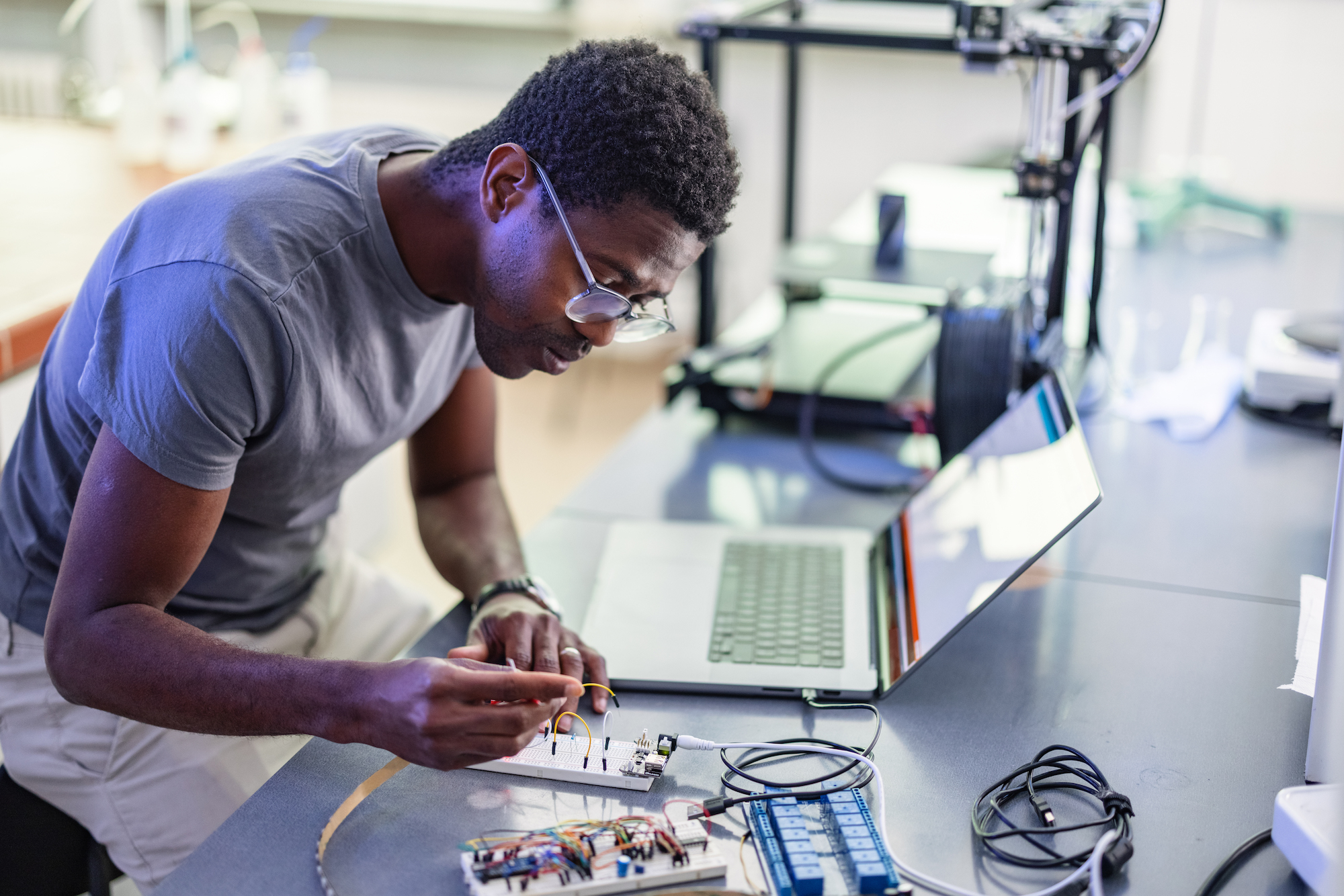 Young African-American technician sitting in the chair, thinking and repairing his 3D printer in the laboratory.